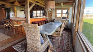 A rustic dining area with a long table and wicker chairs, a bar area with stools, and a view of the outdoors through large windows. - Susie’s Place - Cozy Cabin (Bozeman)