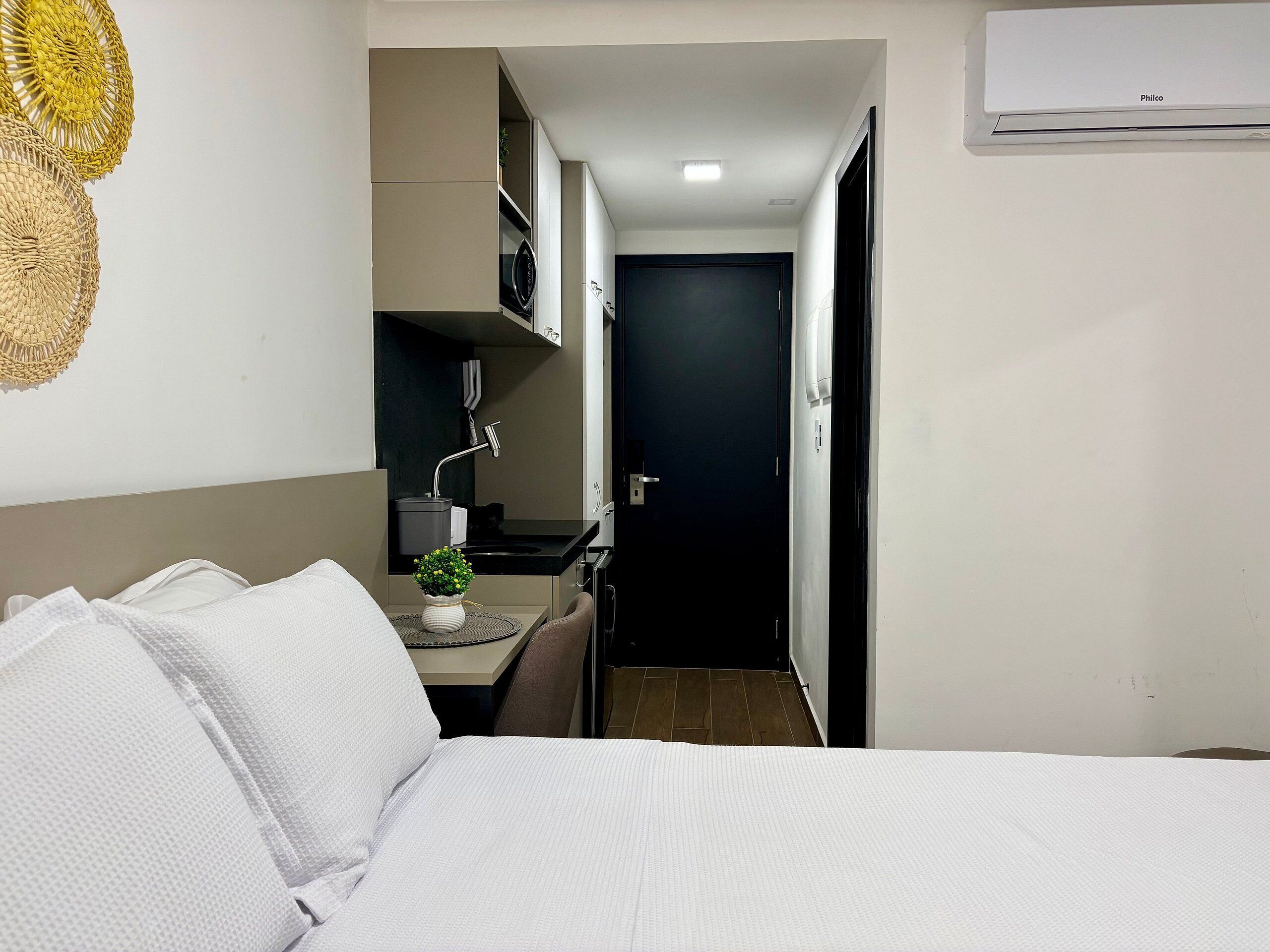A hotel room with a bed, a small desk with a plant, a kitchenette, and a dark door.