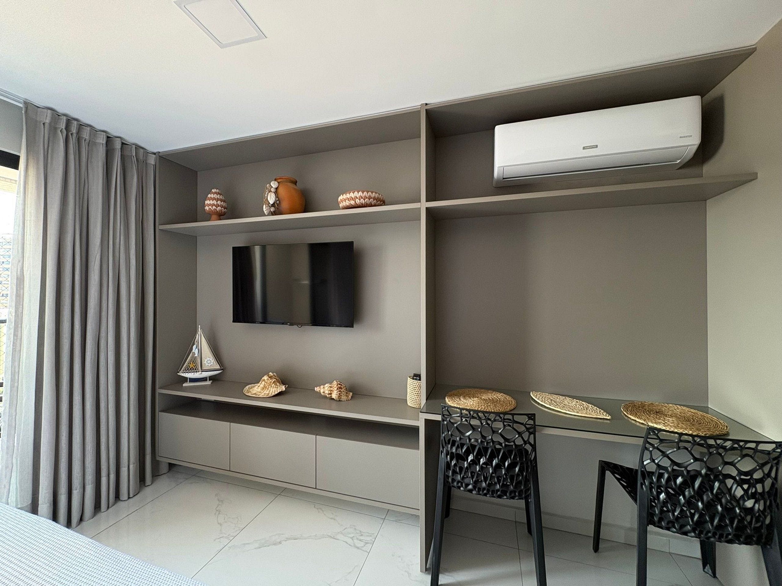 A modern living room with a flat-screen TV, a dining table with chairs, and a built-in air conditioning unit.