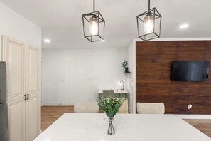 Une cuisine moderne avec des meubles blancs, une table à manger agrémentée d’un vase de fleurs et un mur en bois servant de décor, sur lequel est fixée une télévision.