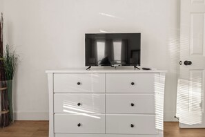 Une commode blanche sur laquelle est posée une télévision à écran plat, une porte dotée d’une poignée et une fenêtre avec des stores.