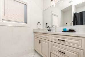 Une salle de bain avec un meuble-lavabo blanc, un miroir et une fenêtre avec des stores.