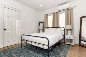 Une chambre à coucher comprenant un lit, une table de chevet, une lampe, un miroir, une porte et un appareil de climatisation.