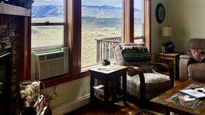 Un salon chaleureux avec une cheminée, une grande fenêtre en arc de cercle offrant une vue sur les montagnes, un fauteuil confortable et une petite table.