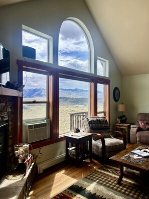 A cozy living room with a fireplace, a large arched window offering a mountain view, a comfortable chair, and a small table.