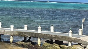 Vista costera con un muelle de hormigón que se adentra en el agua, sostenido por rocas.
