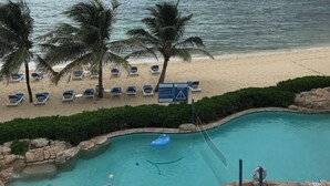 Vista de playa con piscina, palmeras y sillas de descanso.