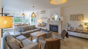 Un salon spacieux avec un grand canapé d’angle, une table basse et une cheminée. On y trouve également des luminaires suspendus et un coin repas avec des chaises.