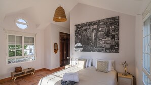 Une chambre avec un grand lit, une table de chevet, une lampe suspendue, une fenêtre avec des stores, un banc en bois et une œuvre d’art représentant un paysage urbain accrochée au mur.