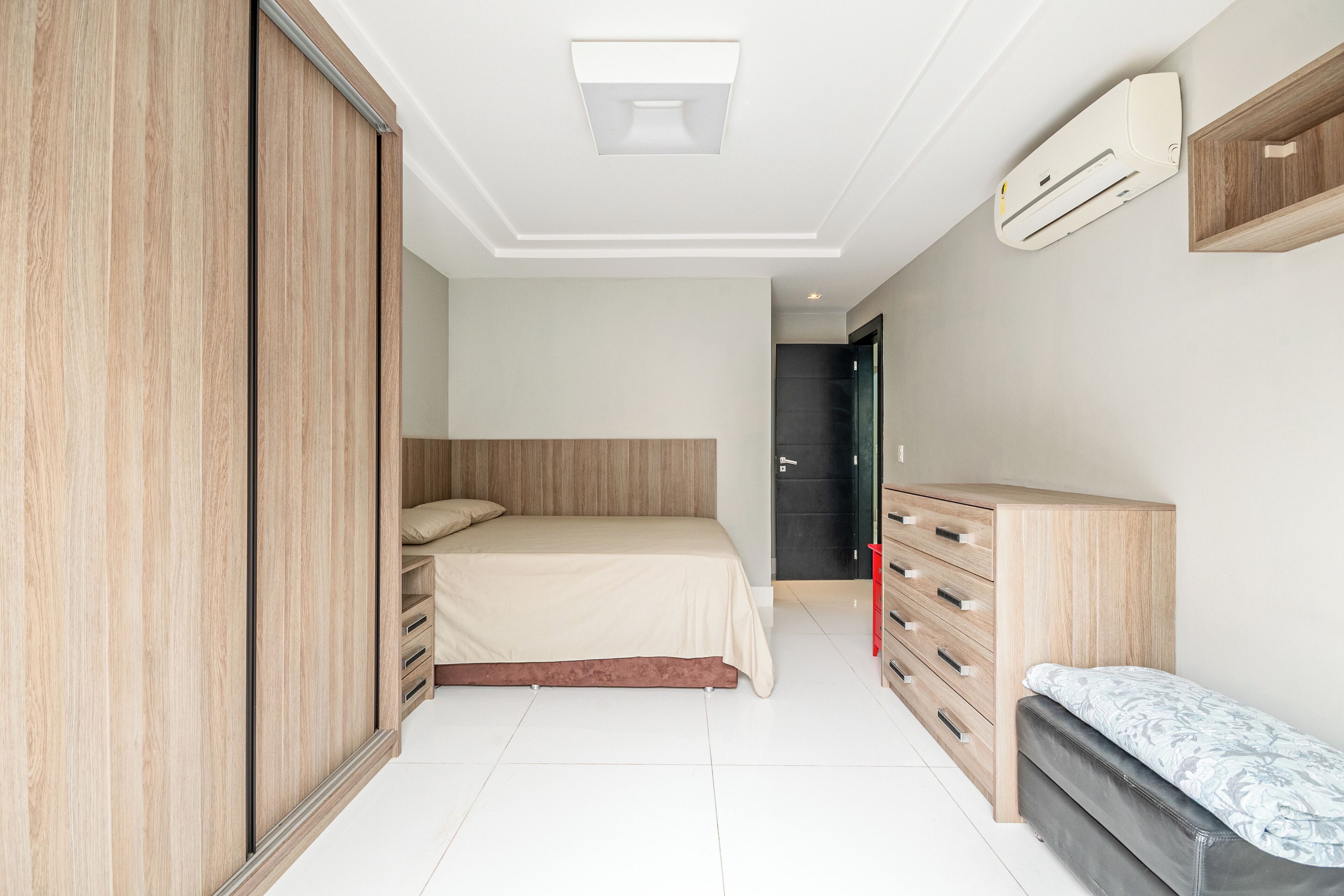 A bedroom with a bed, wooden wardrobe, a bench, a dresser, and an air conditioning unit.