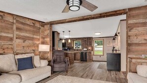 Un interior rústico con pared de madera, un sofá, una silla, un tapiz y una zona de cocina con refrigerador y pileta.