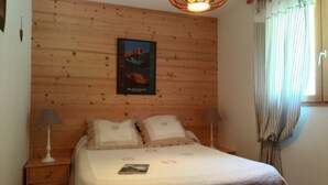 A bedroom with a wooden headboard, two bedside tables with lamps, a bed with a white comforter and pillows, a framed poster on the wall, and a window with curtains.