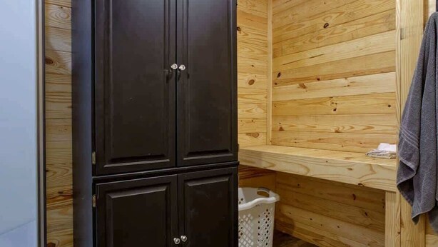 A wooden cabin interior with a dark wooden wardrobe, a wooden bench, and a white laundry basket. - The Lodge at Welch - Both the Big and Little House (Welch)