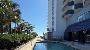 Ein Schwimmbecken mit einer Metallleiter, umgeben von einem Pflasterweg, neben einem mehrstöckigen Gebäude mit Balkonen.