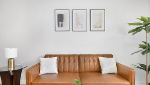 A living room with a brown leather sofa, two white pillows, a glass coffee table, a lamp, and three framed abstract artworks on the wall. - Stylish Queen City Getaway in Camp North End (Charlotte)