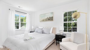 A bedroom with a large bed, a bedside table, a chair, and a window with curtains. - Stylish Queen City Getaway in Camp North End (Charlotte)