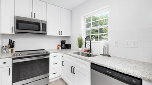 A modern kitchen with white cabinets, stainless steel appliances, a granite countertop, and a window with a view of greenery. - Stylish Queen City Getaway in Camp North End (Charlotte)