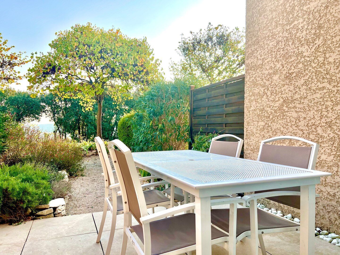 Eine Terrasse mit weißem Tisch und Stühlen, umgeben von Grünpflanzen und Blick auf das Meer.