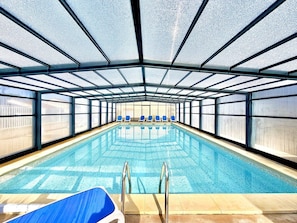 An indoor swimming pool with clear glass walls, blue chairs along the sides, and a metal ladder leading into the pool. - Panorama du Mas, 2 guests Pool in Gard (Calvisson)