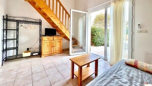 A spacious living area with a wooden staircase, a sliding glass door leading to a balcony, a wooden cabinet with a television, and a bed with a quilt. - Panorama du Mas, 2 guests Pool in Gard (Calvisson)