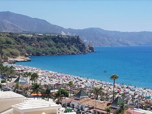 A coastal beach with numerous umbrellas and people, a clear blue sea, and mountainous terrain in the background.