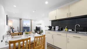 A modern kitchen with white cabinetry, a dining area with a wooden table and chairs, and a view of the city through a large window. - Panoramic City Views – Elegant 2BR, 2 Min to Metro (Salford)