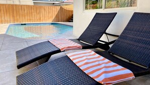 A poolside area with lounge chairs and a view of a wooden fence and a building with large windows. - LUXE Villa 2mi BEACH+BOAT Rental+HTD POOL+SPA! (Fort Lauderdale)