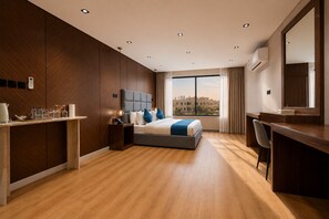 A modern hotel room with a large bed, a desk, a chair, a large window with a city view, and a built-in air conditioning unit.