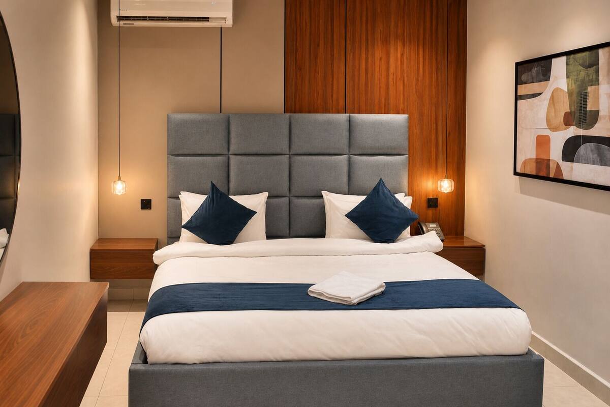 A modern hotel room with a large bed, wooden paneling, a headboard, bedside lamps, and a framed artwork on the wall.