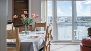 Un comedor con una mesa dispuesta para cuatro, un florero con flores y vistas a un río y edificios a través de amplios ventanales.