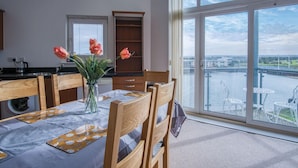 Un comedor con una mesa dispuesta para dos, un florero con flores y vistas a un río y edificios a través de amplios ventanales.