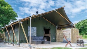 A rustic cabin with a covered porch, a small outdoor seating area, and a visible chimney. - Paviland - Safari Glamping Tent - Llangennith (Swansea)
