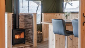 A cozy room with a brick fireplace, wooden walls, and a bar area with two black chairs. - Paviland - Safari Glamping Tent - Llangennith (Swansea)