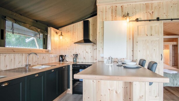 A compact kitchen with wooden cabinetry, a sink, a stove, and a dining area with chairs. - Paviland - Safari Glamping Tent - Llangennith (Swansea)