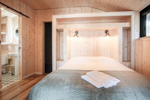A wooden cabin room with a bed, two wall-mounted lights, and a visible bathroom with a toilet and sink.
