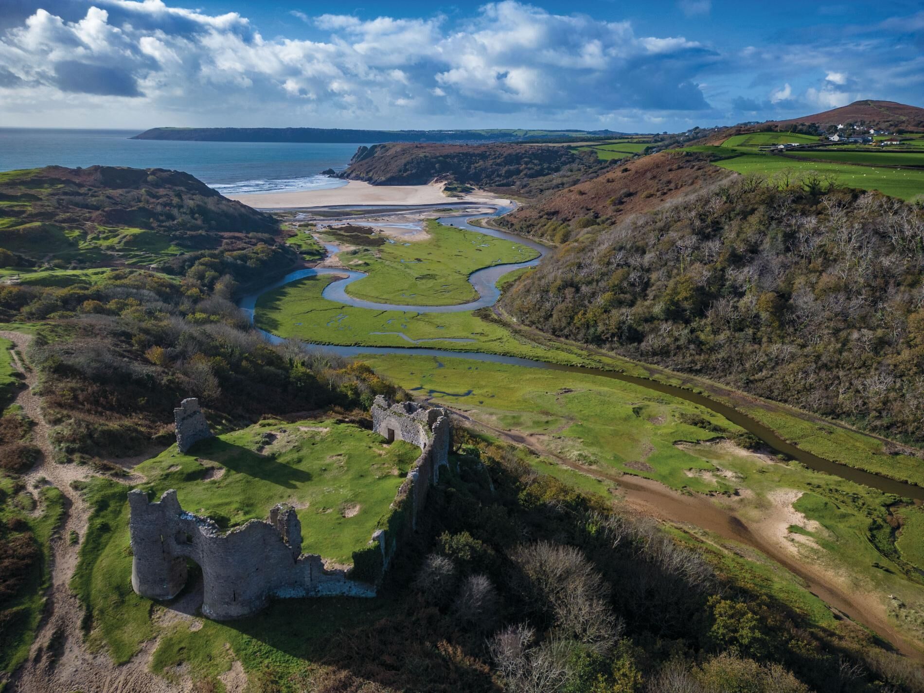 An aerial view of a coastal landscape with a castle ruin, a winding river, and a beach.