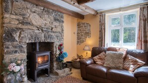 A cozy living room with a stone fireplace, leather sofa, and a large window with curtains. - Rose Cottage - 3 Bedroom Cottage - Parkmill (Swansea)