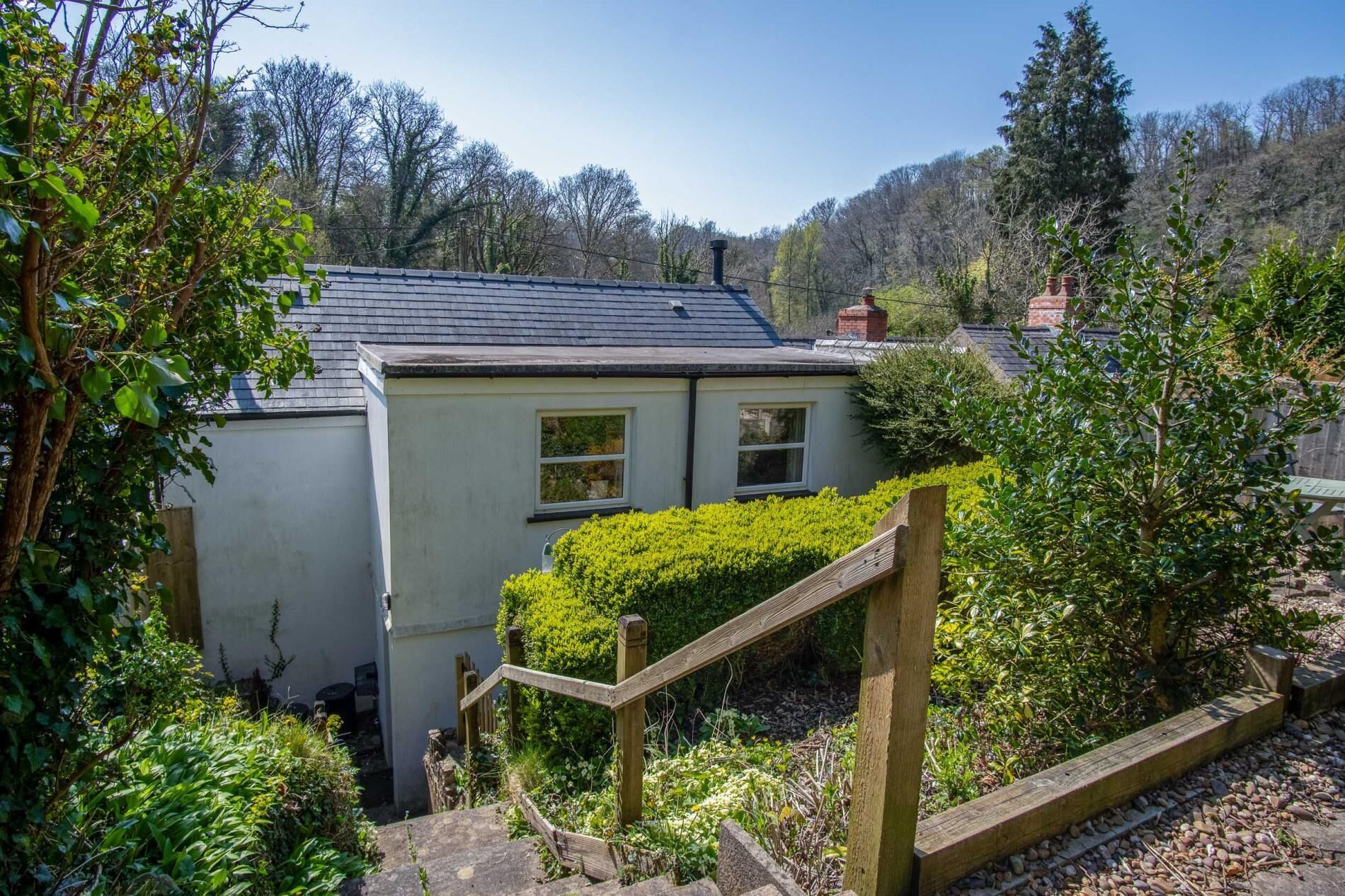 A small, single-story house with a sloped roof, surrounded by lush greenery and trees.