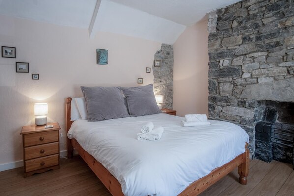 A bedroom with a stone wall, a wooden bed, a nightstand, and a lamp. - Rose Cottage - 3 Bedroom Cottage - Parkmill (Swansea)