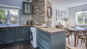 A modern kitchen with a stone accent wall, a built-in washing machine, and a dining area with a view. - Taf Fechan - 3 Bedroom House - Pontsticill (Merthyr Tydfil)