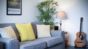 A grey sofa with patterned cushions, a lamp, a guitar, and a framed picture of Brecon Beacons National Park. - Cefn Road - 3 Bedroom Bungalow - Glais (Swansea)