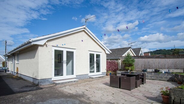 A small, single-story building with a white exterior and a flat roof. It features a large glass door and a small window. There is a paved patio area with outdoor furniture and potted plants. - Cefn Road - 3 Bedroom Bungalow - Glais (Swansea)