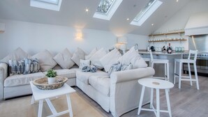Un loft moderno con techo alto, tragaluces, una zona de estar acogedora con sofá y sillones, una pequeña mesa redonda y una cocina con estufa y estanterías.