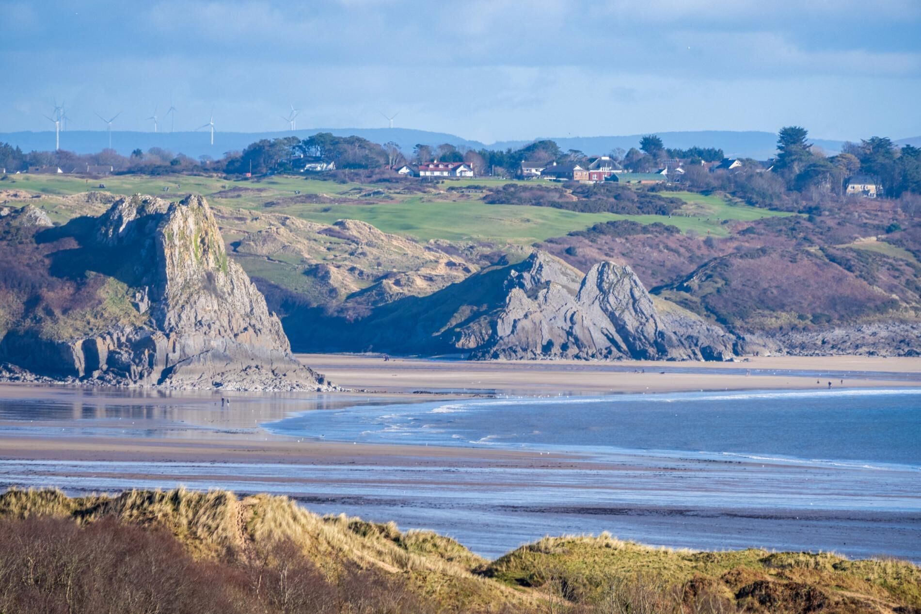 Paisaje costero con acantilados rocosos, una playa y casas al fondo.