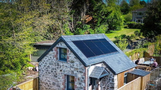 Un edificio de piedra con techo inclinado y paneles solares, rodeado por una cerca de madera y follaje.