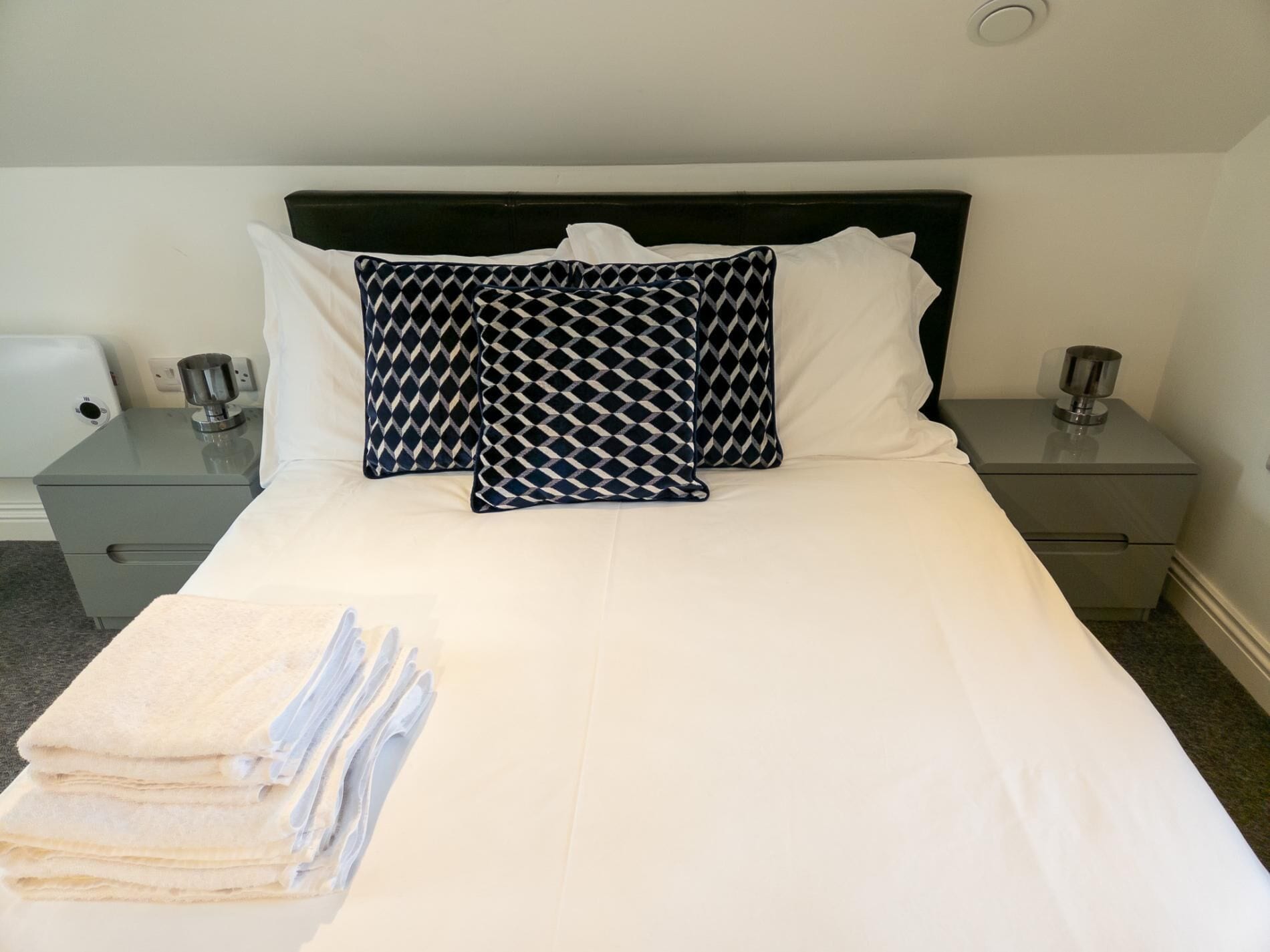 Habitación de hotel con una cama bien hecha, dos mesitas de noche con lámparas y una pila de toallas sobre la cama.