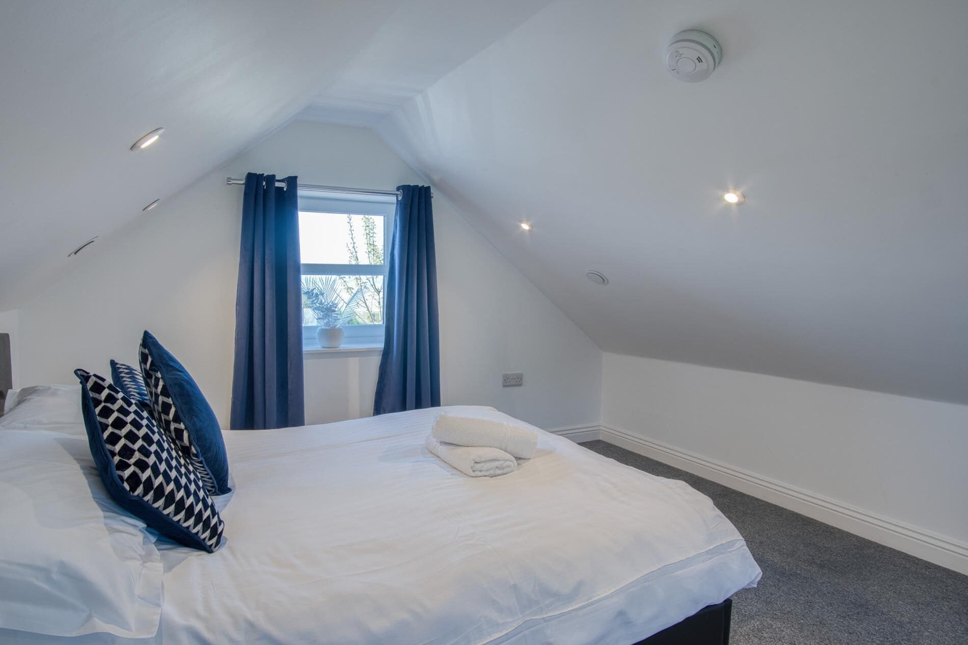 A bedroom with a sloped ceiling, a bed with white linens, blue and black patterned pillows, a window with blue curtains, and a ceiling light.
