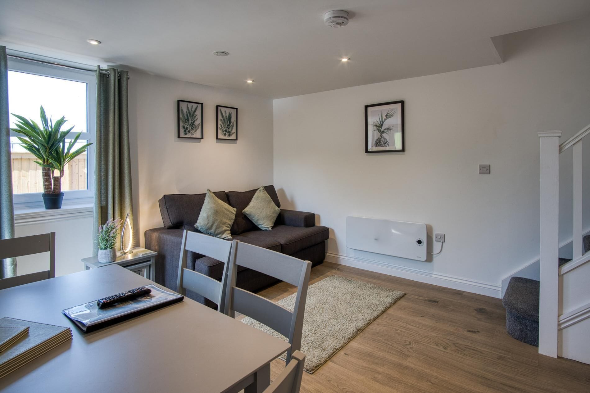 A modern living room with a sofa, a dining table, and a TV. There are framed pictures on the wall and a plant by the window.