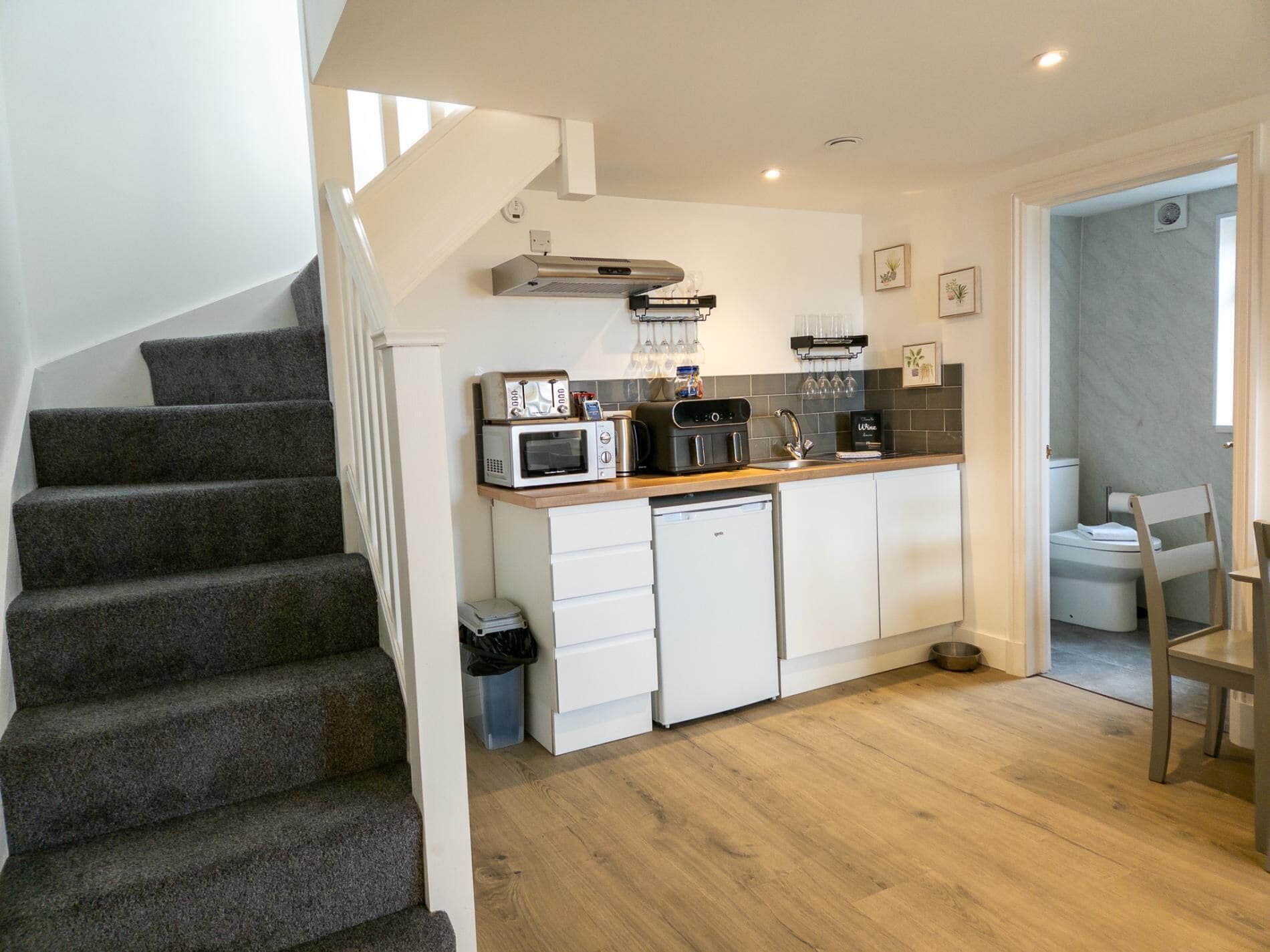 A compact kitchen with a microwave, toaster, and coffee maker on the countertop, a dishwasher, and a small sink. There is a wooden staircase on the left and a door leading to a bathroom.