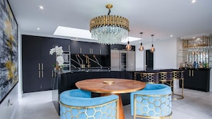 A modern kitchen with black cabinets, a marble countertop, and a central island with a wooden top. There are two blue upholstered chairs with gold accents and a chandelier above.
